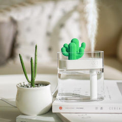 Compact ultrasonic air humidifier releasing fine mist with green cactus design on clear water tank next to small cactus plant on table