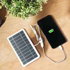Outdoor portable mini solar charger panel connected to a smartphone on a wooden surface next to a small plant