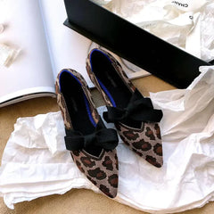 Pair of flat casual boat shoes with pointed toe, leopard print fabric, and black bow detail placed on white paper near an open black shoe box.