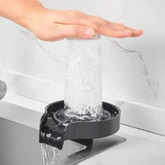 Automatic glass cup washer in black with high-pressure water jet cleaning a glass cup under a hand on white kitchen sink with marble background