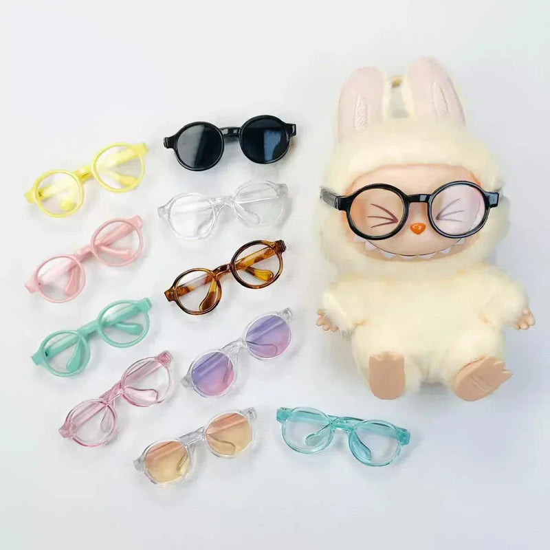 Set of colorful transparent plastic sunglasses for 15cm Labubu dolls including yellow, black, pink, clear, brown, mint, purple, blue shades with a plush doll wearing black round glasses.