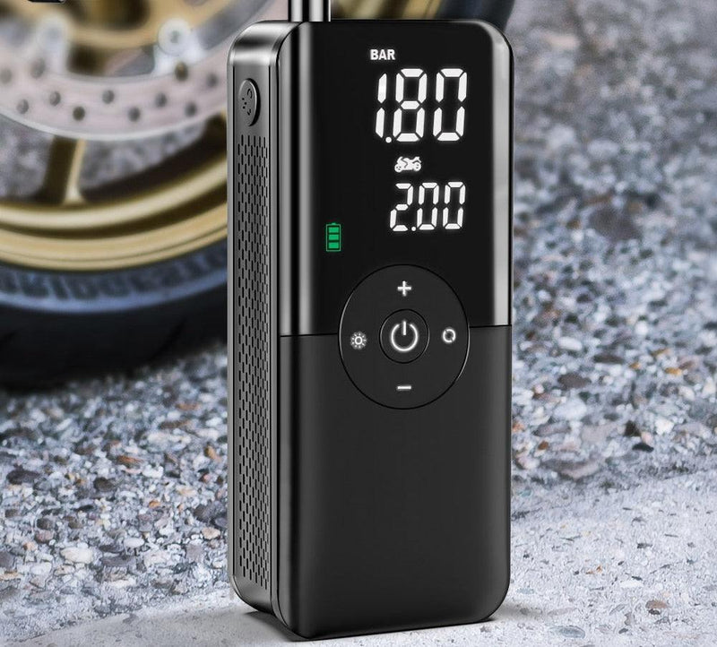 Compact black portable inflator pump with digital pressure display and control buttons next to a motorcycle tire on gravel.