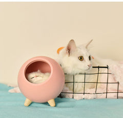 Pink spherical LED cat-shaped light with wooden legs on blue surface next to a white cat in a wire basket